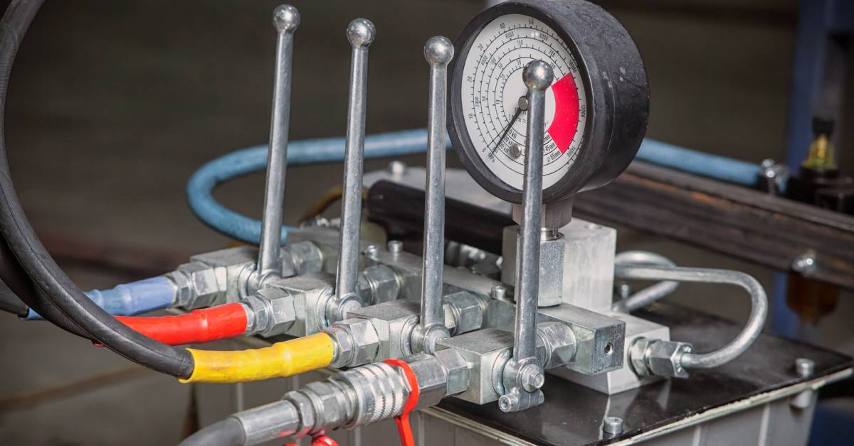 A close-up of a hydraulic system control panel. A pressure gauge, lever, and hoses are coming from the panel.