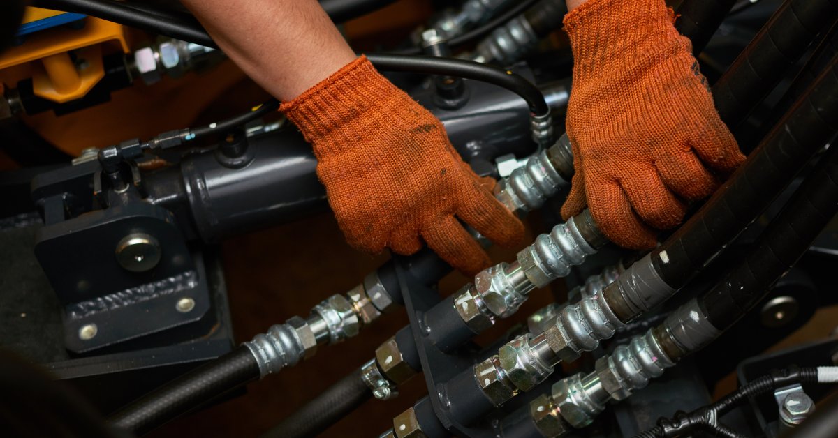 Hands wearing orange gloves perform maintenance on a hydraulic system. The gloves are dirty and worn.