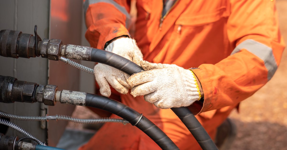 A worker in an orange safety jacket and wearing dirty gloves is checking on a hydraulic hose line. Another hose line is next to it.