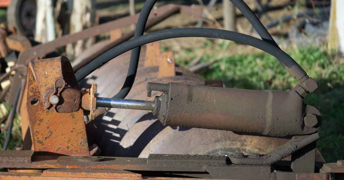 A component of a hydraulic system that is very rusted rests in a field. Other farming equipment is in the background.