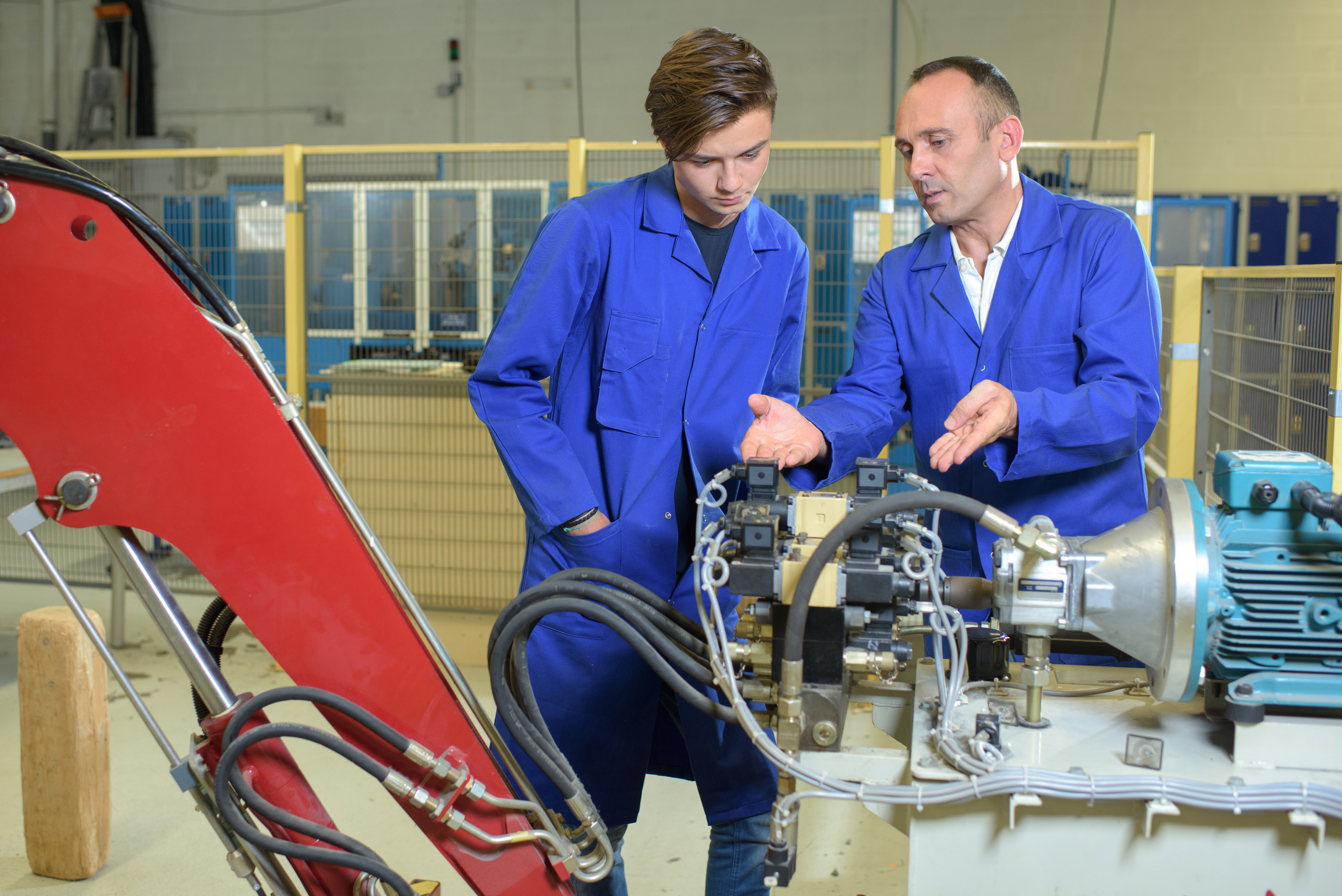 Two white male engineers wearing blue jumpsuits look at a hydraulic arm mechanism, while one guy has his hands out.