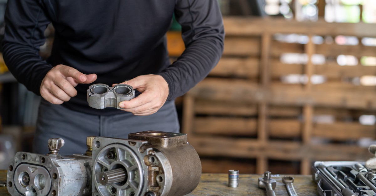 A man is holding a component of a hydraulic pump. The pump and an assortment of tools are on the table in front of him.