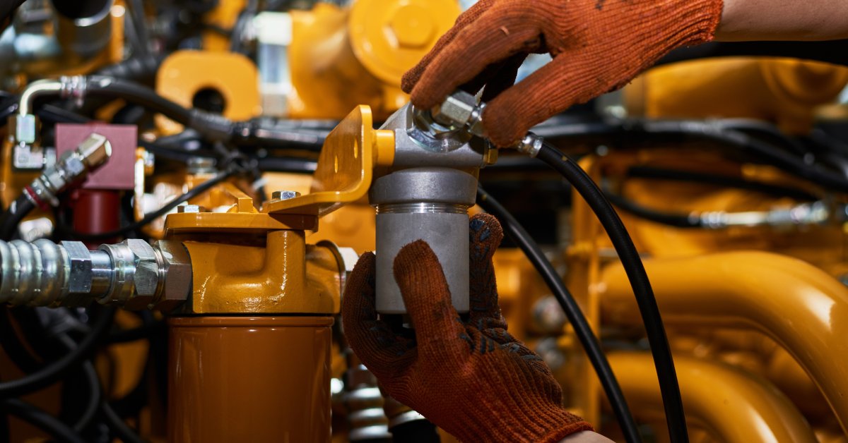 Someone wearing dirty orange maintenance gloves holds a component of a hydraulic system. The system is coated in orange paint.