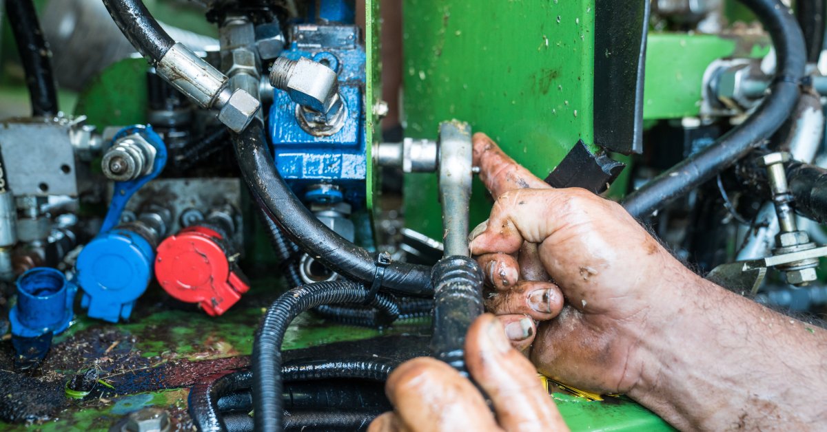 A hand uses a tool to repair a component on a hydraulic system. There is grease on the hands, and the equipment looks dirty.