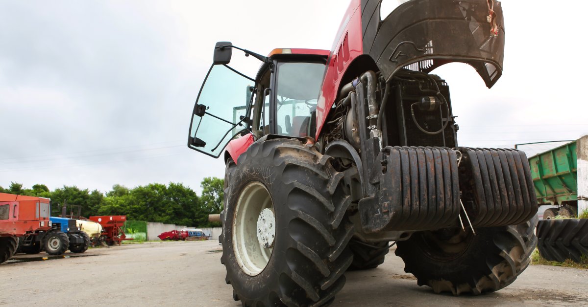 An agriculture machine is parked in a lot with its hood open. Other equipment and vehicles are in the background.